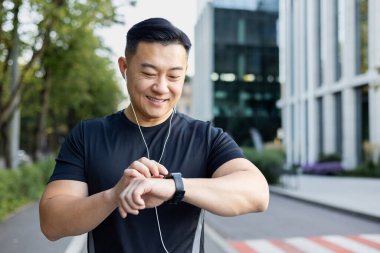 Yakın plan fotoğraf. Genç Asyalı erkek sporcu şehir caddesinde duruyor kulaklık takıyor ve saate bakıyor..