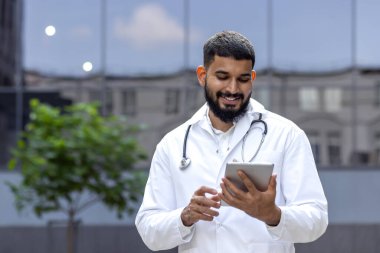 Gülümseyen genç Arap doktor kliniğin yakınındaki caddede duruyor ve tablet kullanıyor..