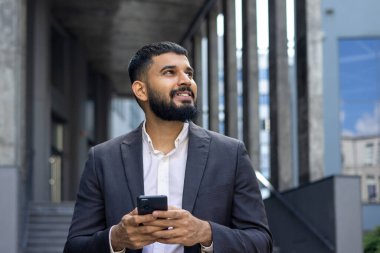 Başarılı genç Müslüman bir erkek işadamı bir ofis merkezinin yanında duruyor, cep telefonu kullanıyor ve gülümsüyor. Yakın plan fotoğraf..