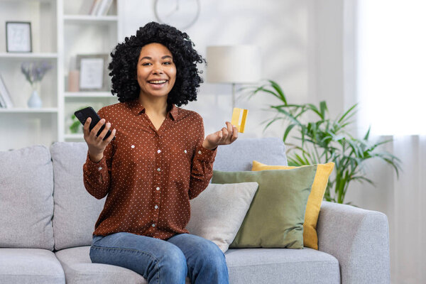 Portrait of a smiling young African American woman sitting on the couch at home, holding a credit card and phone, smiling at the camera.