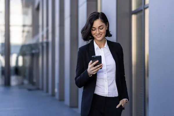 A young beautiful business woman is walking outside the office building, a woman in business clothes is smiling, using an application on the phone, browsing the Internet, typing a message.