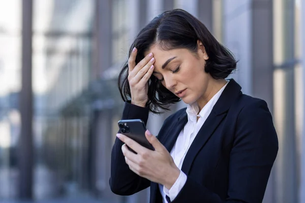 Businesswoman received notification message with bad news, woman reading online on phone, upset and confused sad using smartphone app, outside office building, walking in business suit.