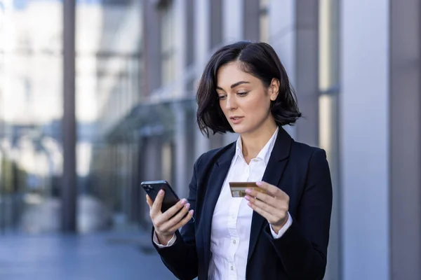 A young business woman is standing on the street near the work office and using a credit card and a phone.