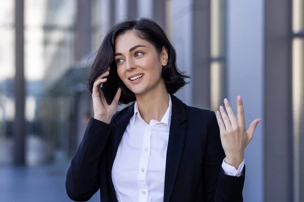 Young successful satisfied business woman talking on the phone, female worker in a business suit walking around the city outside an office building.