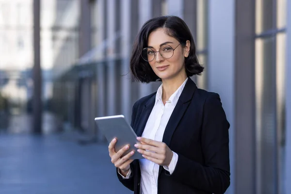 Portrait of a self-confident business woman, executive director, company founder standing in a suit and glasses near the office center, holding a tablet in her hands and smiling at the camera.