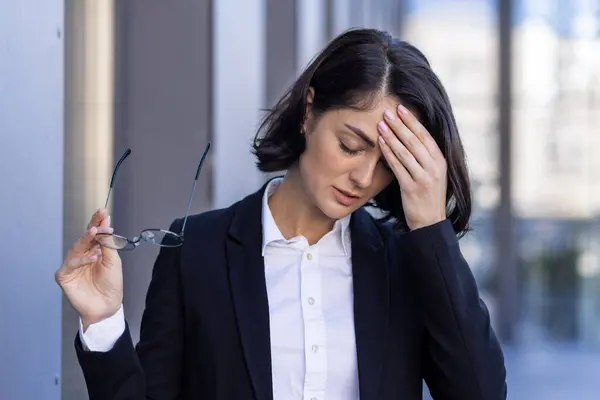 Young woman tired with headache, businesswoman outside office building rubs eyes, dizzy, overworked worker outdoors in business suit close up.