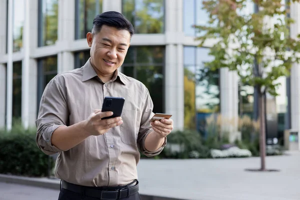 A young smiling Asian man is standing on a city street, holding a credit card and using a mobile phone.