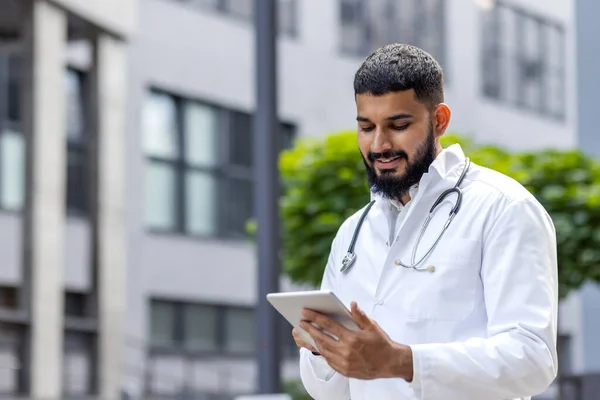 A young Muslim doctor in a bedlam robe stands outside the hospital and uses a tablet, writes, types, consults.
