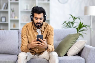 Kulaklıklı genç Hintli bir adam elinde bir telefon tutuyor, parlak bir oturma odasında bir kanepeye oturuyor, müzik dinliyor, dinlenmekten hoşlanıyor..