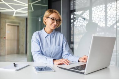 Ofisteki masada oturan, dizüstü bilgisayarda çalışan, gülümseyen ve ekrana bakan dost canlısı sarışın işçi..
