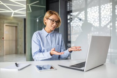 Gözlüklü bir kadın, dizüstü bilgisayarın önünde bir masaya oturur, video aracılığıyla iletişim kurar, müşterilere tavsiyelerde bulunur, bir sürü jest yapar..