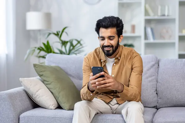 A happy Indian man is sitting on the sofa in the living room, holding a smartphone, smiling and looking at the screen.