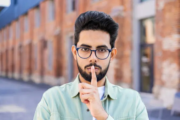 A young Indian man stands on the street, puts his index finger to his lips, looks at the camera.