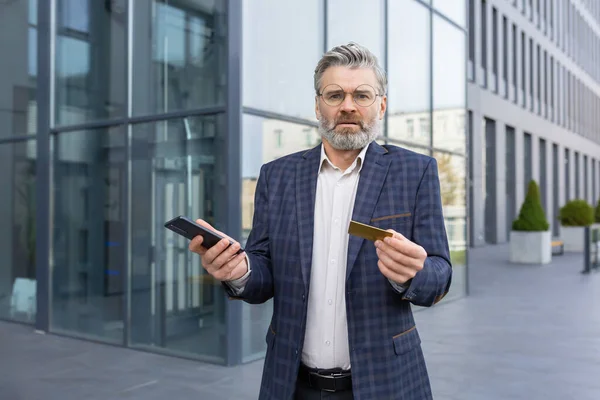 Angry businessman holding a card and a smartphone, standing near an office building, unsuccessful online shopping, fraud on the Internet.