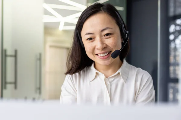Asian female online customer support customer service worker at workplace inside office, woman using headset phone for video call, businesswoman typing on laptop keyboard.