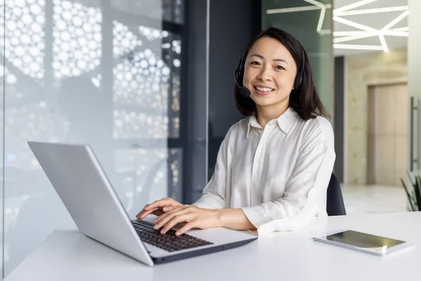 Portrait of online support worker, asian woman with headset phone smiling and looking at camera, businesswoman working in laptop at workplace inside office, call center call center support service.