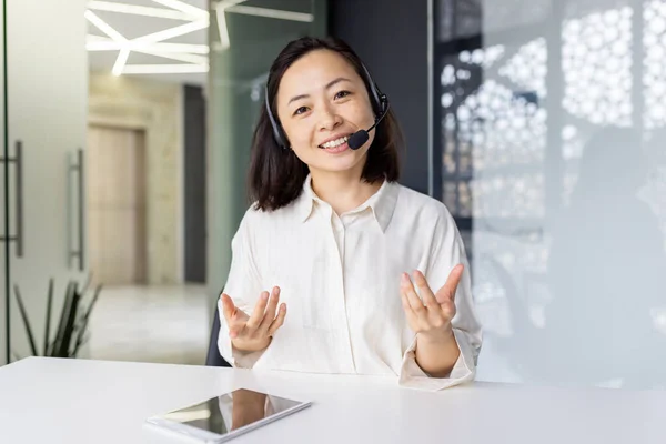 Video call online meet, Asian woman with headset smiling and looking at camera, chatting with smile, online customer service customer service worker at workplace, webcam view.
