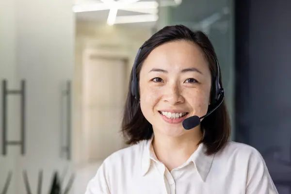 Close-up portrait of Asian online support worker, woman smiling and looking at camera using headset phone for online consultation and video call, working inside office with laptop.