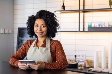 Küçük bir kafe sahibinin portre garsonu. Önlüklü kadın gülümseyip elinde tablet bilgisayarla kameraya bakıyor. Kahve tezgahının yanındaki bir restoranın çalışanı..