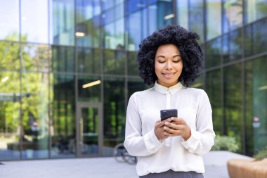 Genç ve güzel bir Afro-Amerikalı kadın ofis binasının önünde yürüyor, iş kadını elinde bir telefon tutuyor, mutlu bir şekilde gülümsüyor, internette geziniyor ve akıllı telefonuyla başvuru yapıyor..