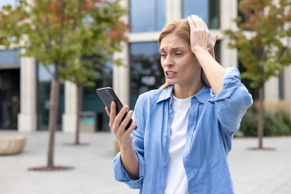 Mature woman outside office building in blue shirt sad received online message, female worker reading bad news using app on phone, viewing social media bullying and harassment.