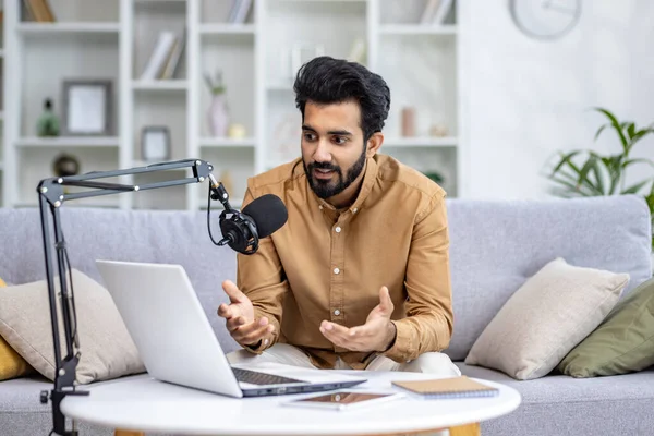 A young Indian man sits at home on the sofa in front of a laptop and speaks into a microphone, records an online podcast, gives an interview, explains by gesturing with his hands.