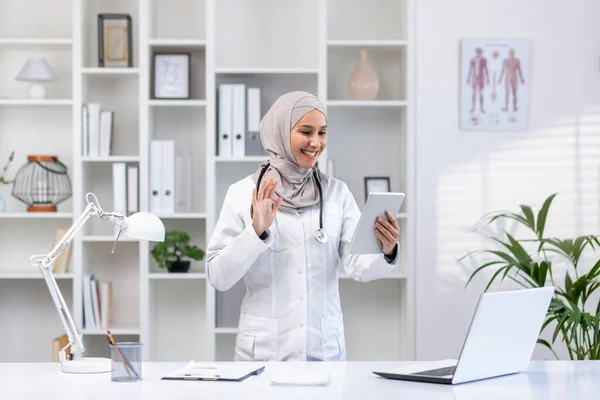 A smiling Muslim doctor in a hijab conducts an online appointment with a patient, holds a tablet, waves at the camera and says hello.