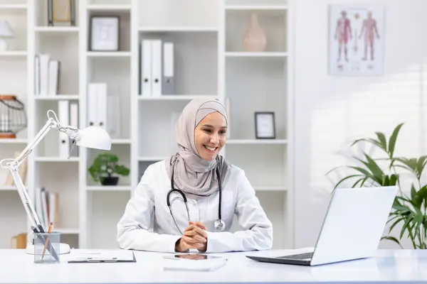 A smiling female doctor at an online appointment, sits at a table in front of a laptop, consults a patient remotely.