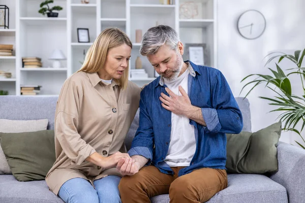 Frightened wife holding hand of husband having heart attack and holding hand to chest, sitting on sofa in living room at home.