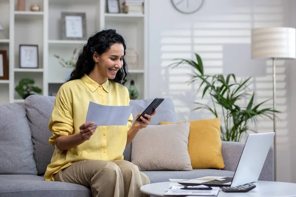 Happy hispanic woman working in home office sitting on sofa in front of laptop, holding phone and documents in hands, remote work.