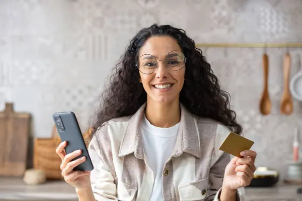 Happy hispanic woman holding phone and card, online shopping, internet payment, smiling and looking at camera.