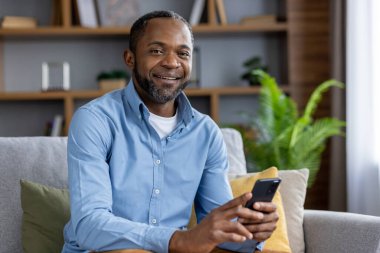 Yetişkin bir adamın portresi, Afro-Amerikan bir adam gülümseyen ve elinde telefonla kameraya bakan, evdeki koltukta oturmaktan mutlu, akıllı telefondaki uygulamayı kullanarak.