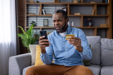 Üzgün ve üzgün bir adam oturma odasında kanepede oturuyor, son sınıf öğrencisi Afro-Amerikan 'ı aldatıyor telefon ve banka kartından memnun değil, fon aktarım hatası, hesap bloğu.