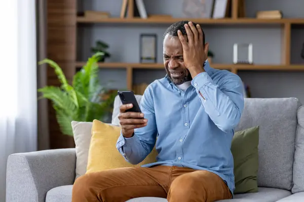Sad nervous man sitting at home on sofa in living room of house, African American man received bad news online, read news on social media, received negative feedback via email.