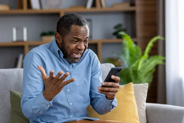 Sad nervous man sitting at home on sofa in living room of house, African American man received bad news online, read news on social media, received negative feedback via email.