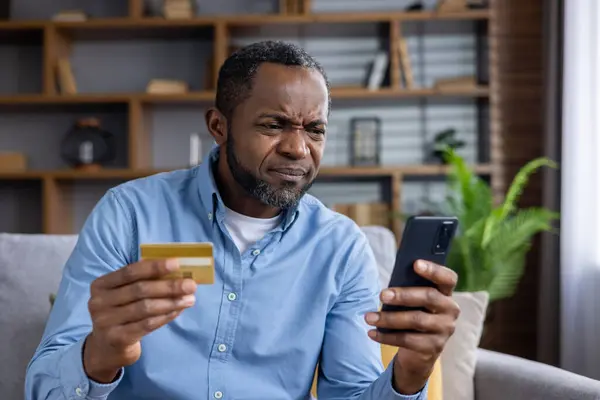 Upset and sad man sitting at home on couch in living room, cheated senior mature african american man displeased holding phone and bank credit card, rejected fund transfer error, account block.