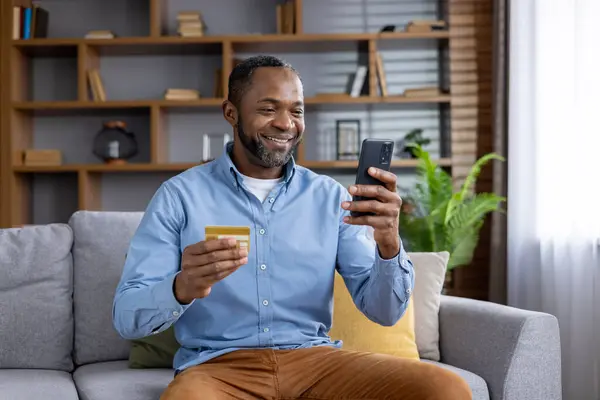 Joyful mature man at home on sofa with phone and bank credit debit card in hands, satisfied african american man in living room chooses gifts in online store, uses app on smartphone, transfers money.