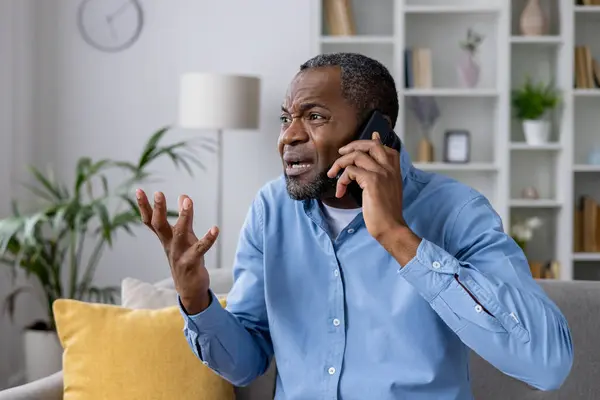 Angry nervous man sitting on sofa at home talking on the phone, unhappy african american man yelling at interlocutor, frustrated user and online shopper.