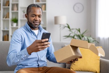 Mutlu ve memnun bir alıcı, bir online mağaza için hediye teslimatı hızıyla tatmin olmuş bir karton kutu aldı, bir Afrikalı Amerikalı adam elinde telefon ile malların alındığını doğruladı.