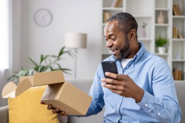 Mutlu ve memnun bir alıcı, bir online mağaza için hediye teslimatı hızıyla tatmin olmuş bir karton kutu aldı, bir Afrikalı Amerikalı adam elinde telefon ile malların alındığını doğruladı.