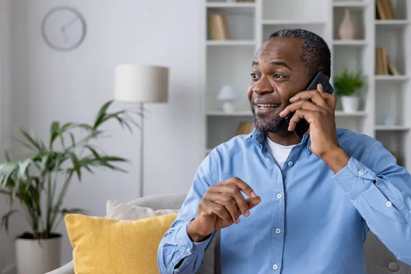 Happy and satisfied man talking on the phone sitting at home on the sofa in the living room, mature senior African American man in the house smiling telling the interlocutor.