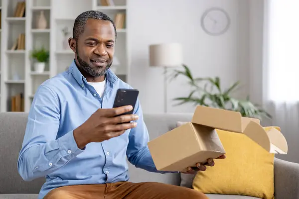 Happy and satisfied buyer received cardboard box in mail, satisfied with speed of delivery of a gift for an online store, an African American man with a phone in hands confirms receipt of the goods.