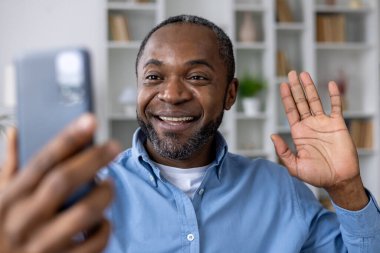 Telefondaki uygulamayı video görüşmesi için kullanan mutlu adam, Afro-Amerikan adam gülümseyen ve akıllı telefon kamerasına bakan, evde oturma odasındaki kanepede oturmuş el sallayan adam..