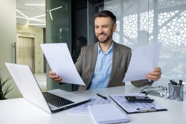 Ofisin içinde başarılı bir finansör portresi. Adam bakıyor, raporları kontrol ediyor, sözleşmeleri elinde tutuyor. İş adamı kılığında gülümsüyor, başarı elde ediyor..