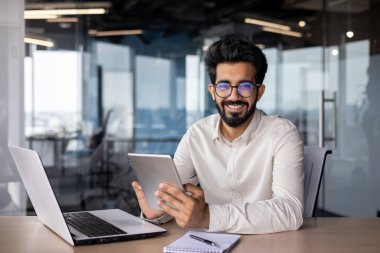 Masada elinde dizüstü bilgisayarla oturup tablet kullanan ve kameraya bakan gülümseyen Müslüman bir adamın portresi..