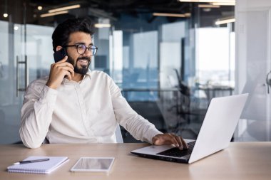 Gülümseyen genç Hintli bir adam ofiste bir masada oturuyor, dizüstü bilgisayar kullanıyor ve telefonla konuşuyor..