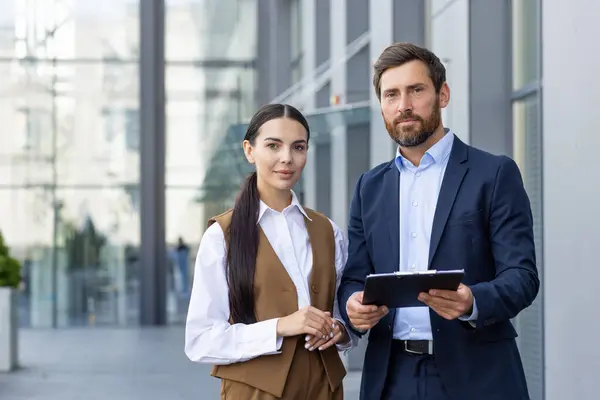 Portrait of a team and partners of a young business man and woman standing outside the office center with documents in their hands, smiling and confidently looking at the camera.