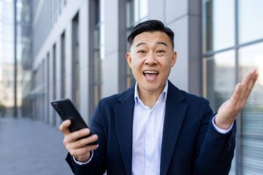 Ofis binasının yanında takım elbiseli Asyalı bir gencin yakın plan portresi. Elinde telefonla kameraya bakıyor. Şokta..