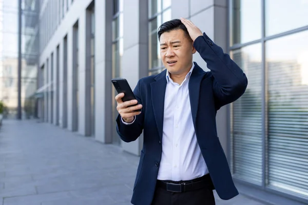 A young Asian man in a business suit is standing outside an office center, looking shocked and worried at the phone screen, and holding his head.