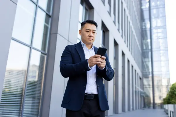 Serious Asian businessman standing on the street near the office with a mobile phone, typing a message, making a call, waiting for an appointment.